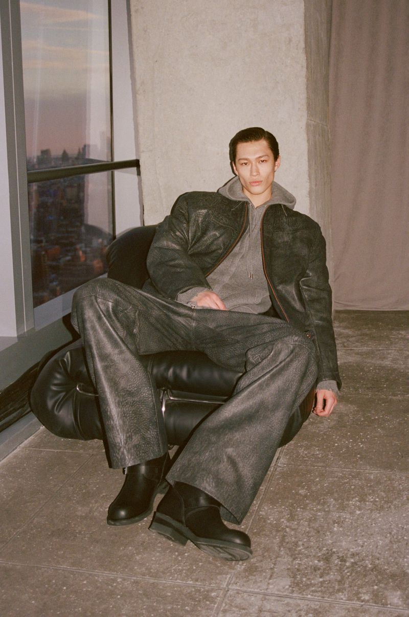 Adult male in a grey hoodie and leather jacket sits in a black chair in an indoor setting near a window with an urban view.