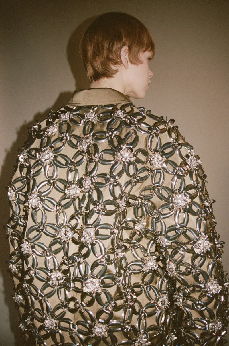 A person from the back wearing a tan and silver jacket with flower embellishments is facing right on a neutral-toned background.