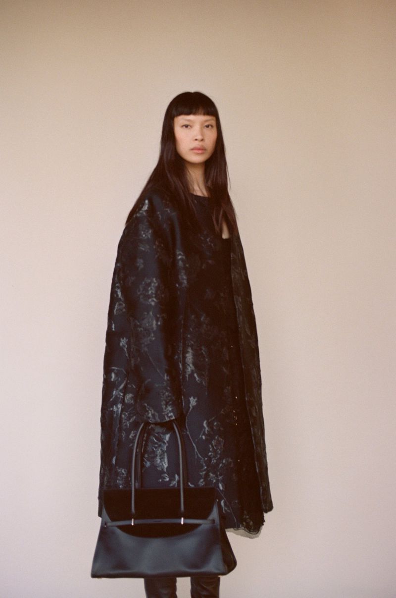 Model Diane Chiu poses in a long dark jacquard coat holding a structured black handbag against a warm neutral studio backdrop.