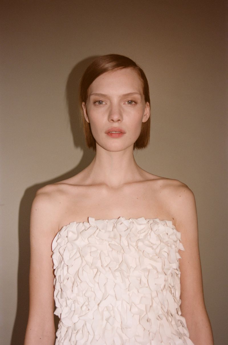 Model with short auburn hair wearing a strapless off-white ruffled textured top against a neutral backdrop.