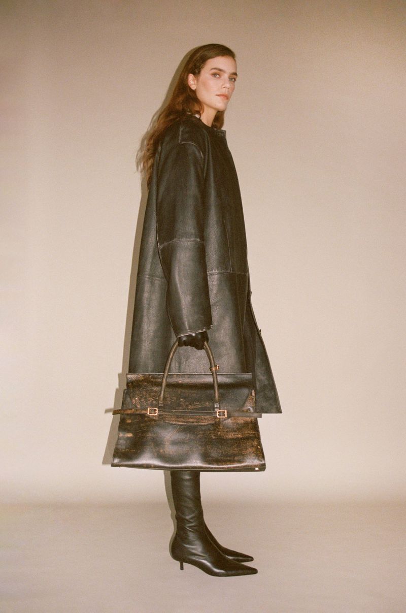 A woman in a long black coat, gloves, and boots stands in front of a beige background, holding a large black leather handbag.