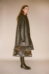 Model in an oversized dark leather coat holding a large textured leather handbag and wearing black pointed-toe boots.