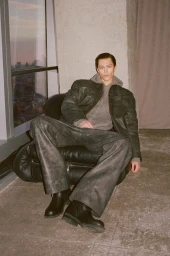 Joshua Thompson wearing a dark jacket, grey hoodie, and wide-leg trousers seated in an armchair for Simkhai Fall 25.