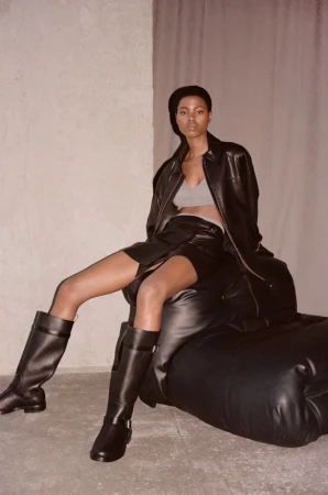 Model seated in a black leather jacket, miniskirt, and tall black leather boots leaning back on a dark leather chair.
