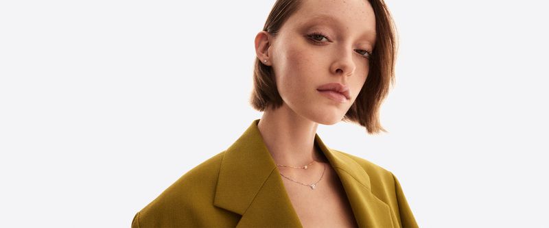 Elegant woman in a mustard-colored blazer with layered necklaces posing in a clean white studio setting, showcasing fashion jewelry.