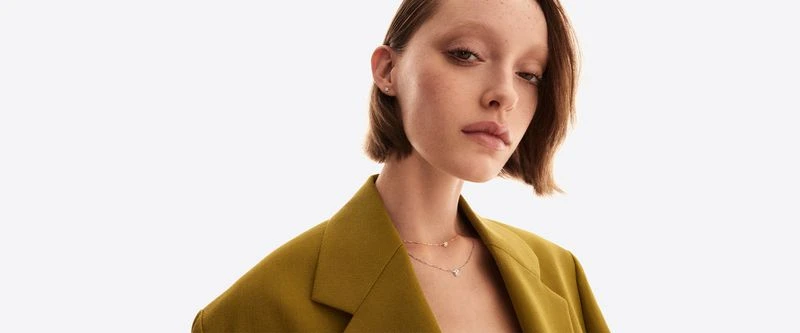 Elegant woman in a mustard-colored blazer with layered necklaces posing in a clean white studio setting, showcasing fashion jewelry.