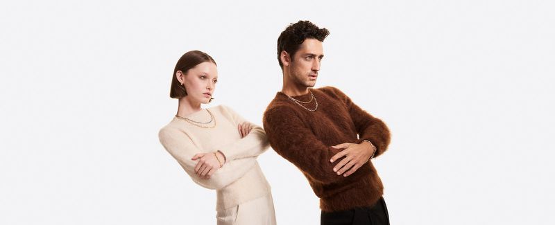 Two fashion models, one man and one woman, pose back-to-back in cozy sweaters and elegant jewelry in a studio setting with soft lighting.