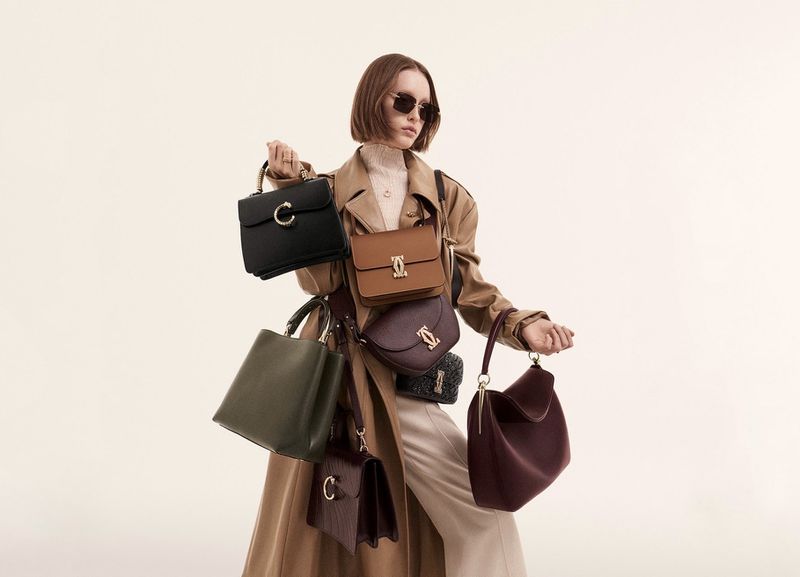 Woman in trench coat poses with multiple handbags in a studio, showcasing various colors, styles, and shapes against a neutral backdrop.