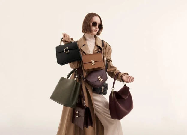 Woman in trench coat poses with multiple handbags in a studio, showcasing various colors, styles, and shapes against a neutral backdrop.