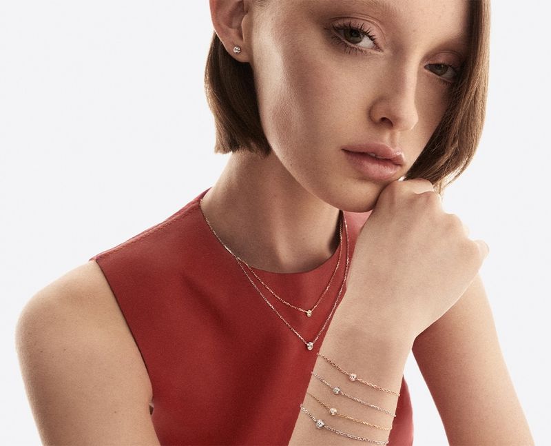 Model Avah Adams wearing layered gold chain necklaces and stacked bracelets in a red top against a white background.