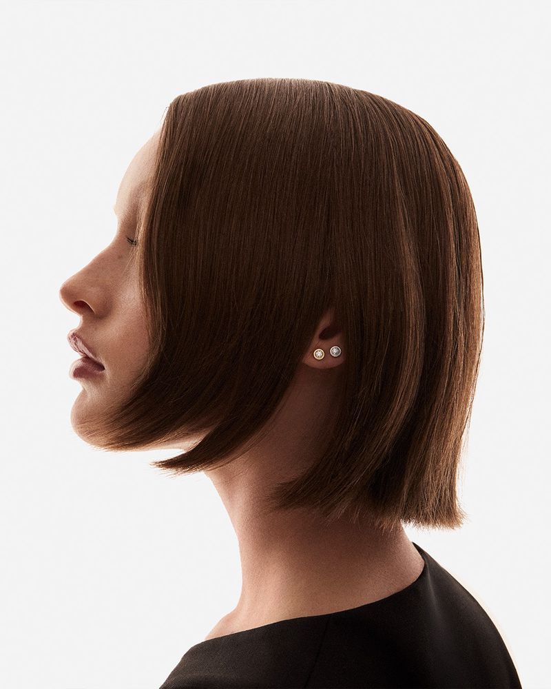 Profile shot of a young fair-skinned adult female with chin-length brown hair, pearl earrings, and wearing a black top against white.