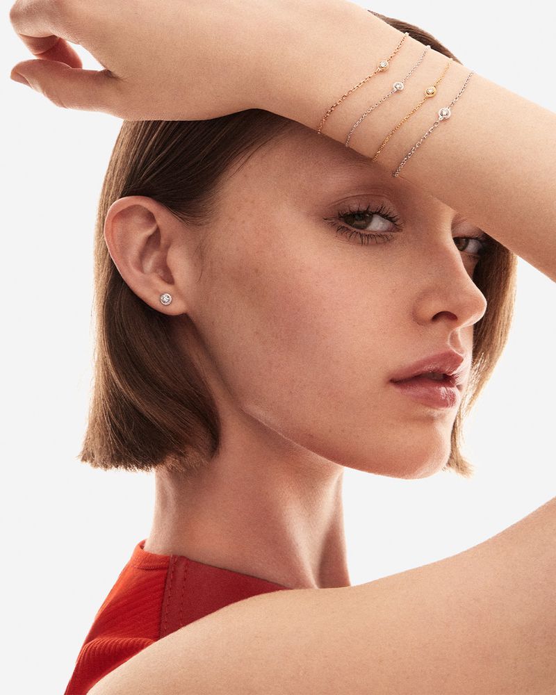 Close-up of a fair-skinned young woman wearing a stud earring with multiple gold chain bracelets around her arm in a light studio.