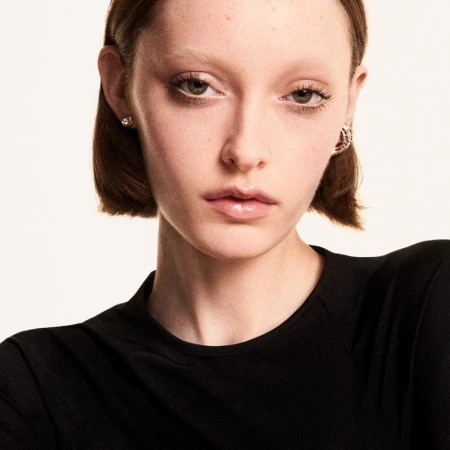 Close-up portrait of model Avah Adams with short brown hair and bleached eyebrows wearing earrings against a white background.
