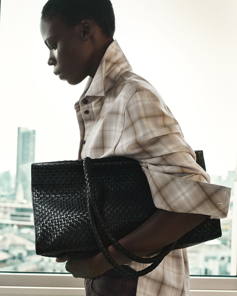 Woman in plaid shirt holding black woven tote bag, standing in front of large window with city buildings in the background.
