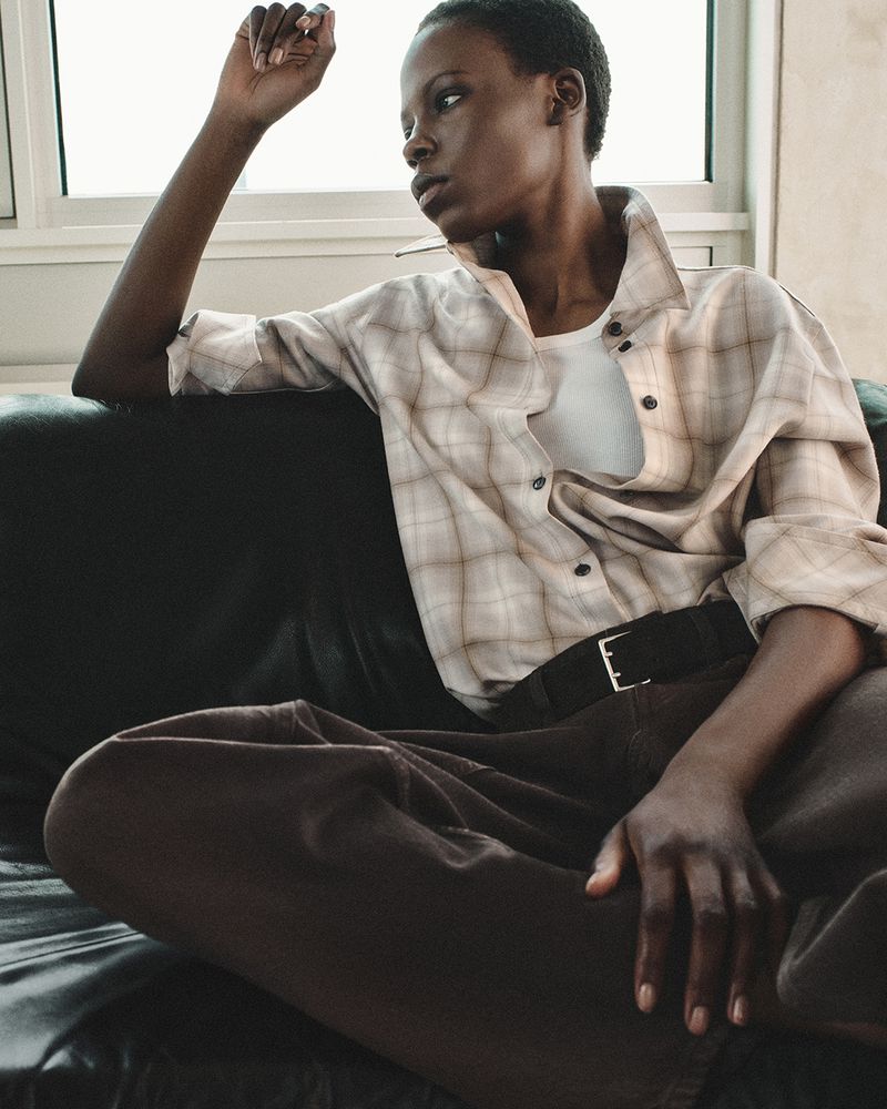 A dark-skinned woman in a plaid shirt and brown pants sits pensively on a dark sofa near a bright window in a casual setting.