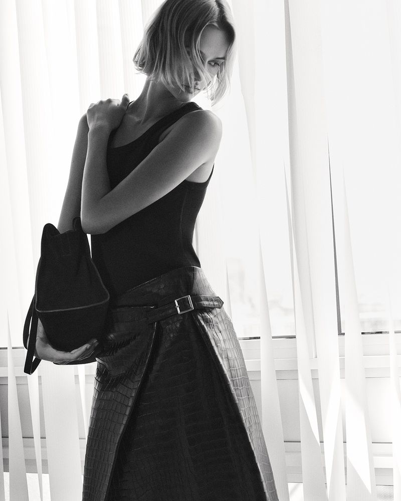 Black-and-white shot of a slender woman with short blonde hair, a dark top, and textured wrap skirt holding a soft tote bag in front of curtains.