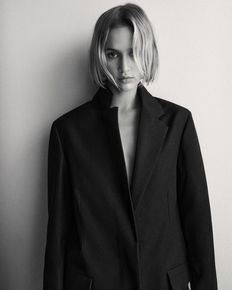 Blonde woman in a black blazer posing against a white wall in a studio, showcasing a blend of fashion and portraiture elements.