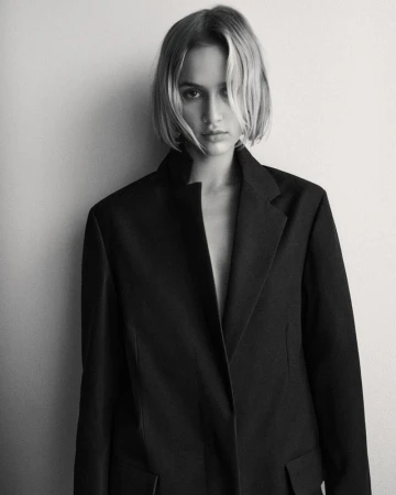 Blonde woman in a black blazer posing against a white wall in a studio, showcasing a blend of fashion and portraiture elements.