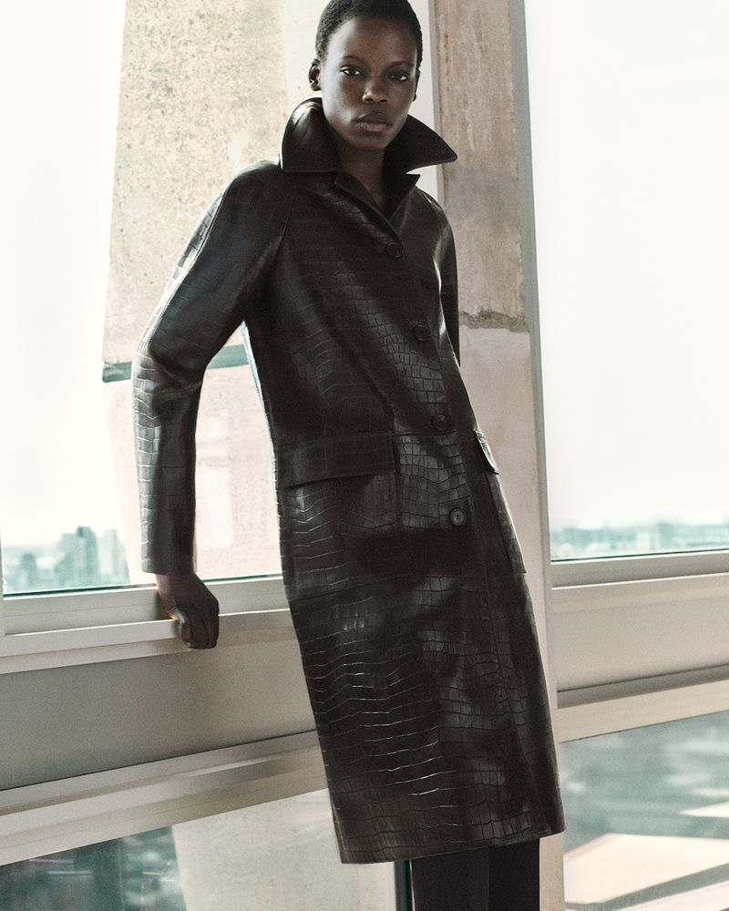Elegant portrait of an African female in a collared dark leather trench coat, standing by a window with an urban view, fashion editorial.