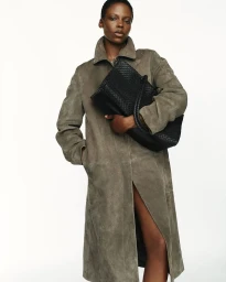 Model Agel Akol wearing a long grey suede coat while holding a large black woven tote bag against a white seamless studio backdrop.