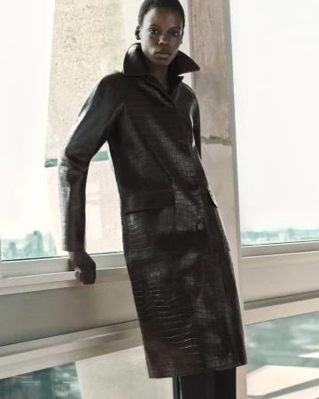 Model wearing a dark textured leather coat standing near a large window for COS Pre-Fall 25 campaign.
