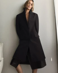 Model Quinn Mora wears a long black tailored coat and embossed leather shorts against a neutral grey studio backdrop for COS.