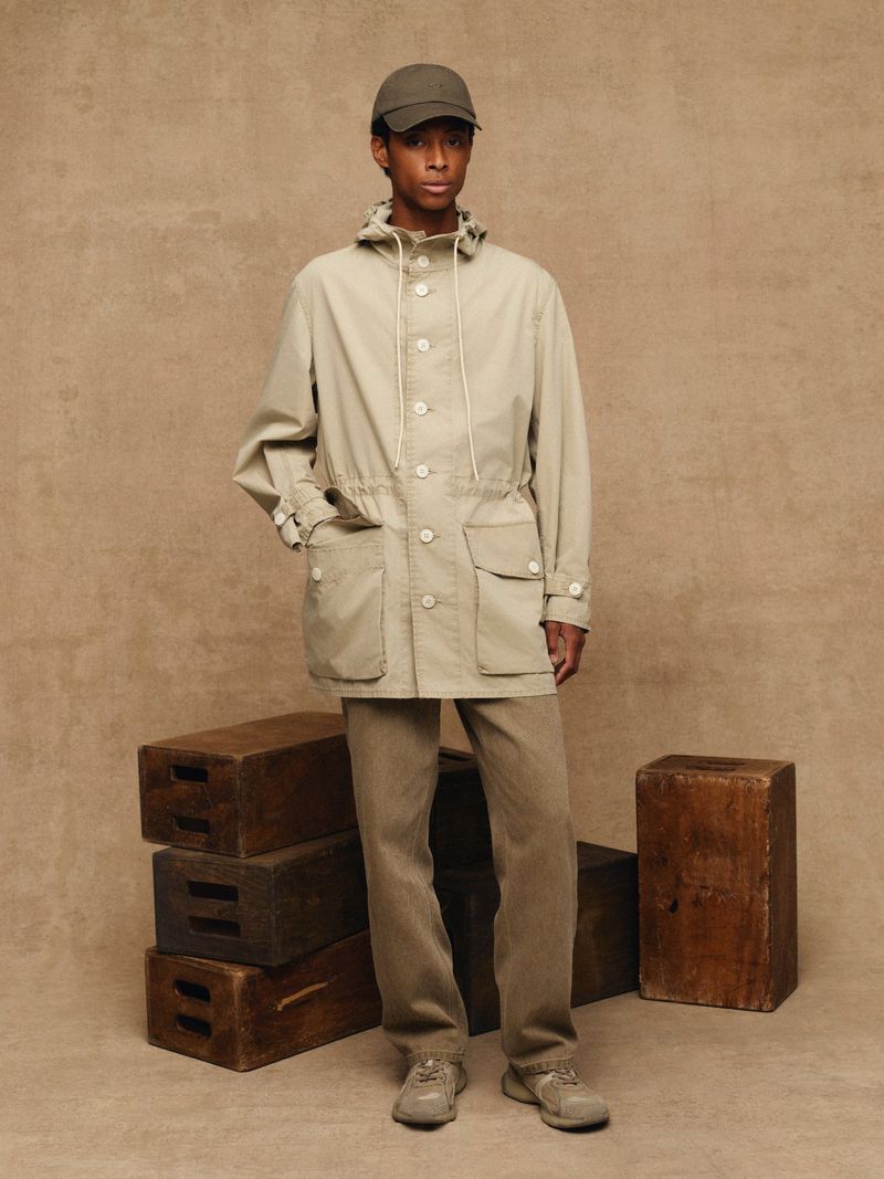 Full-length shot of a young, Black adult male wearing a neutral jacket, pants, and cap, posed in front of a neutral backdrop and wood crates.