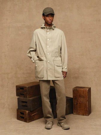 Full-length shot of a young, Black adult male wearing a neutral jacket, pants, and cap, posed in front of a neutral backdrop and wood crates.