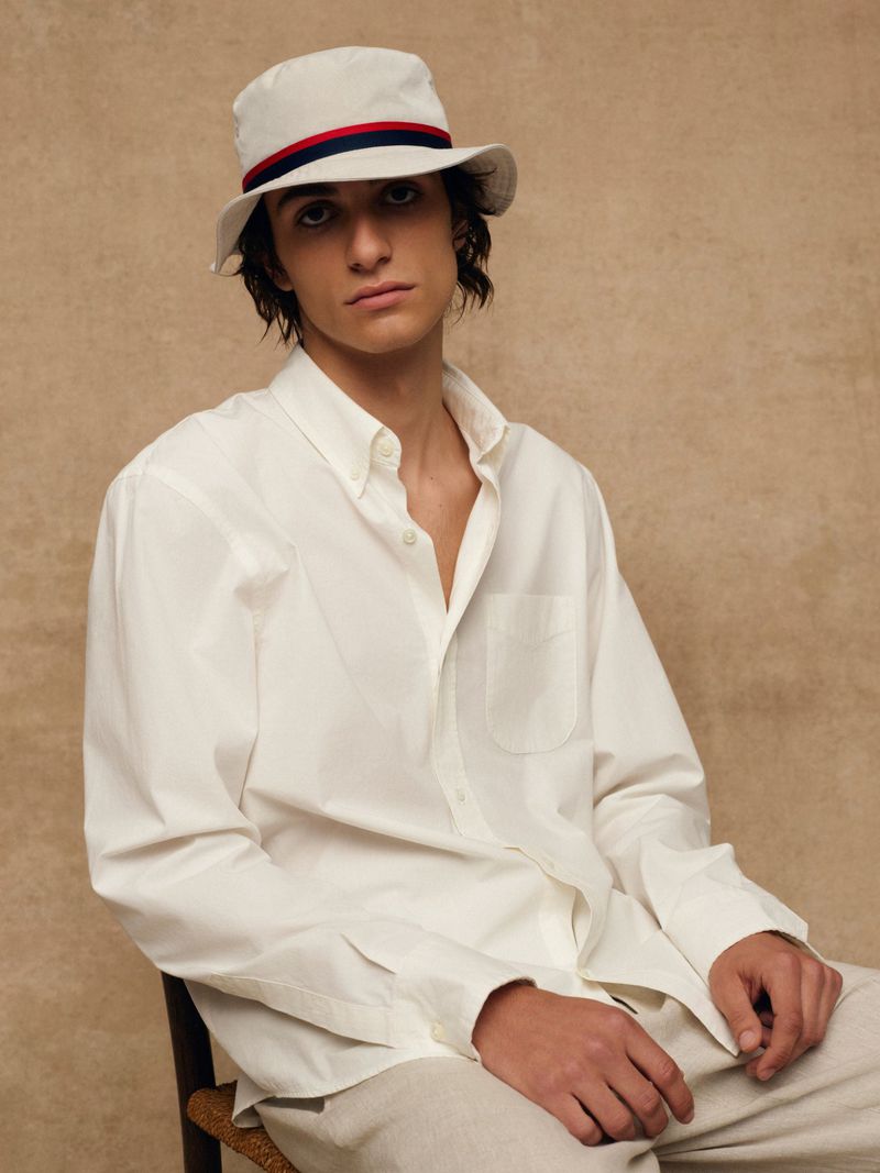 Man in white bucket hat and long-sleeve shirt sitting indoors against a neutral background, looking at the camera with a neutral expression.