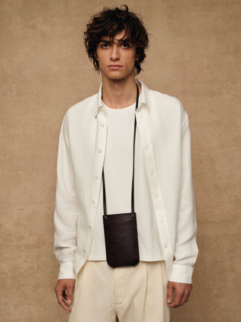 Portrait of a young male adult in a white button-down shirt, white t-shirt, and neutral pants with a dark phone bag.