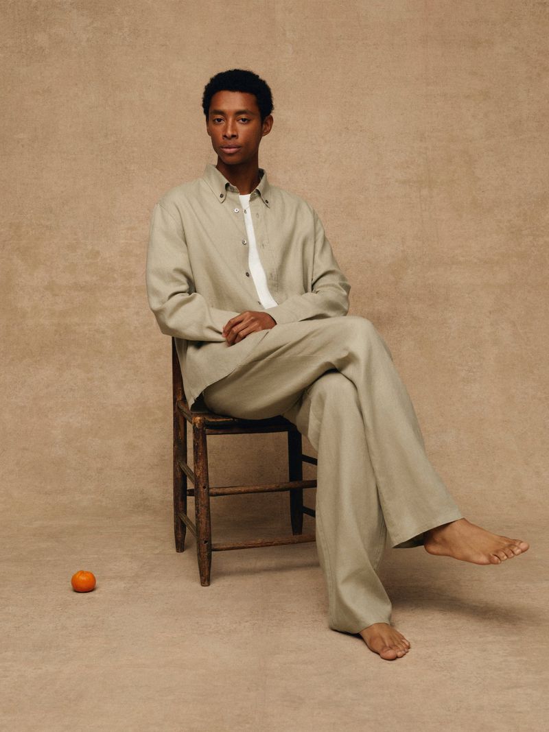 An adult male model is seated on a wooden chair in a studio wearing a sage-colored shirt and matching pants, with bare feet and an orange fruit on the floor.