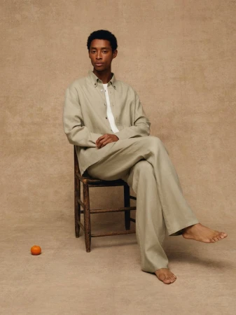 Black man in a sage green matching shirt and trousers seated barefoot on a wooden chair next to an orange on a beige backdrop.