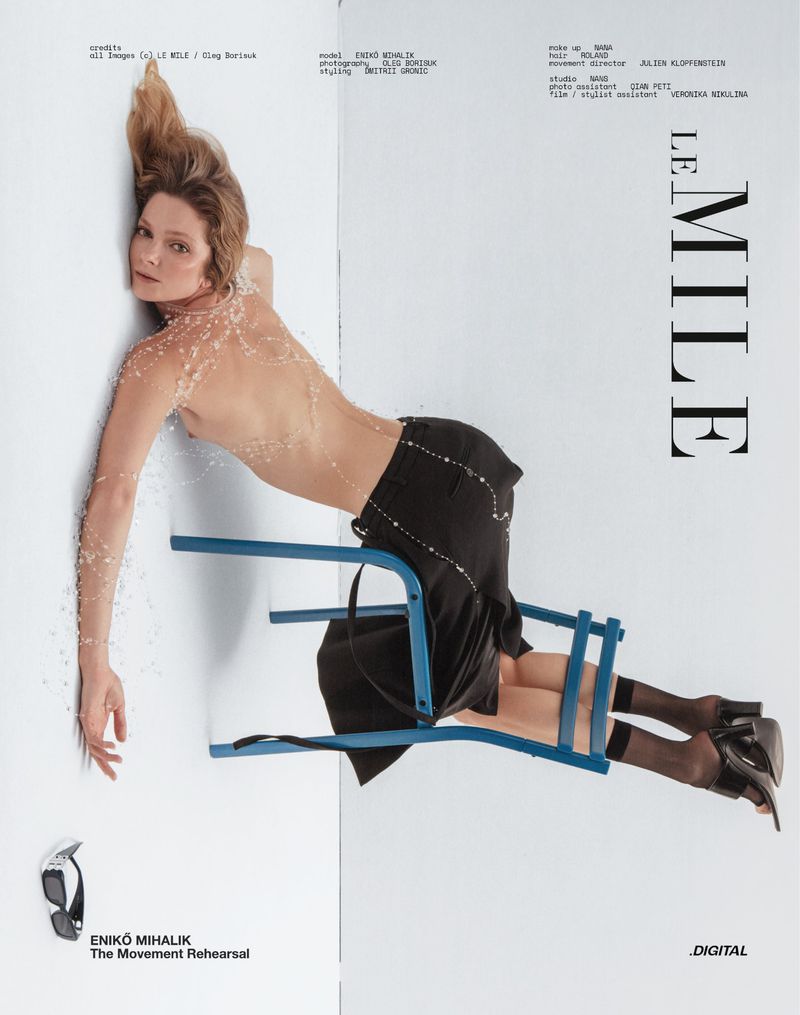 Fashion editorial image featuring Eniko Mihalik lying on a white surface wearing a black skirt and a blue chair. Black high heels are visible.