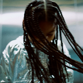 Close-up of a girl with long braided hair, wearing a shiny silver jacket, posing in an indoor environment.