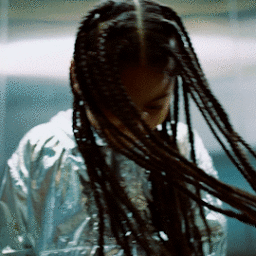 Close-up of a person with long braided hair tossing their head, wearing a shiny silver metallic jacket against an industrial background.