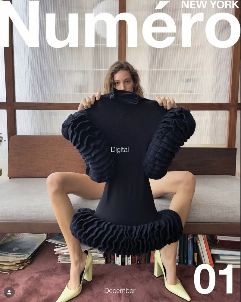 Magazine cover featuring a woman sitting with legs spread holding a black dress, with yellow high heels and books in the background.