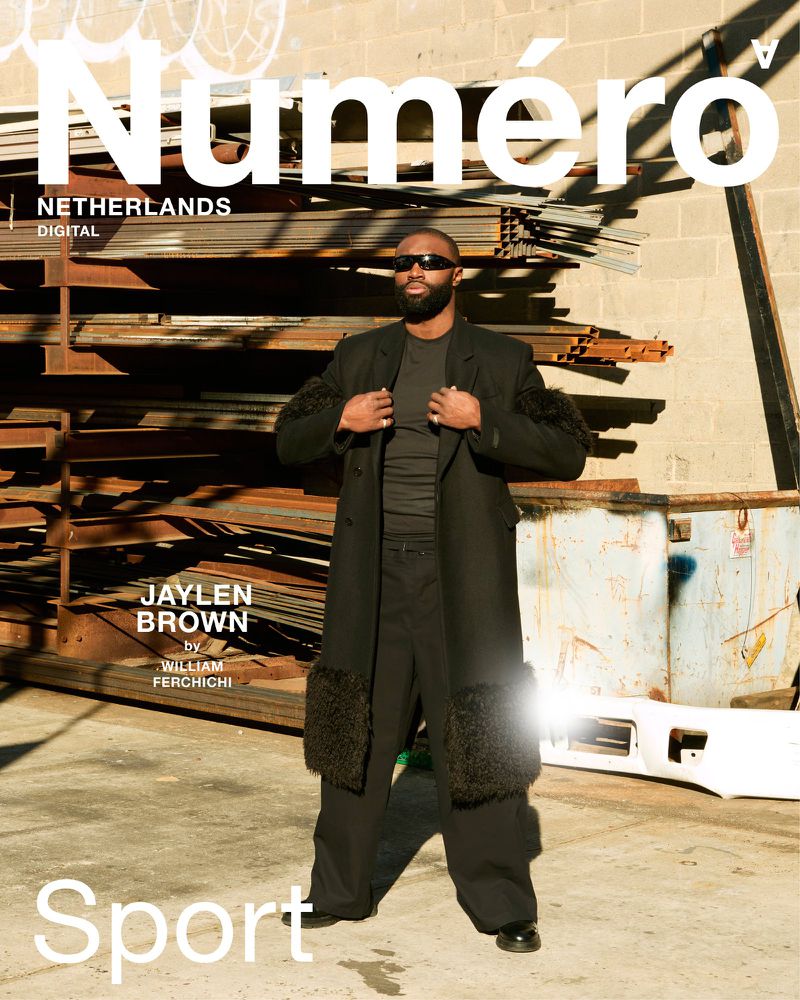 Editorial shot of a Black male model wearing sunglasses and a fur-trimmed coat in an urban setting with metal and concrete background.