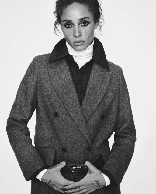 Black and white portrait of a woman with fair skin in a wool coat and a black belt against a bright white background. Fashion editorial.