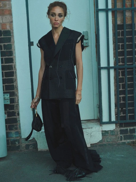 Elegant woman in a black sleeveless blazer and wide-leg pants holds a small purse, posing against a brick wall and doorway backdrop.