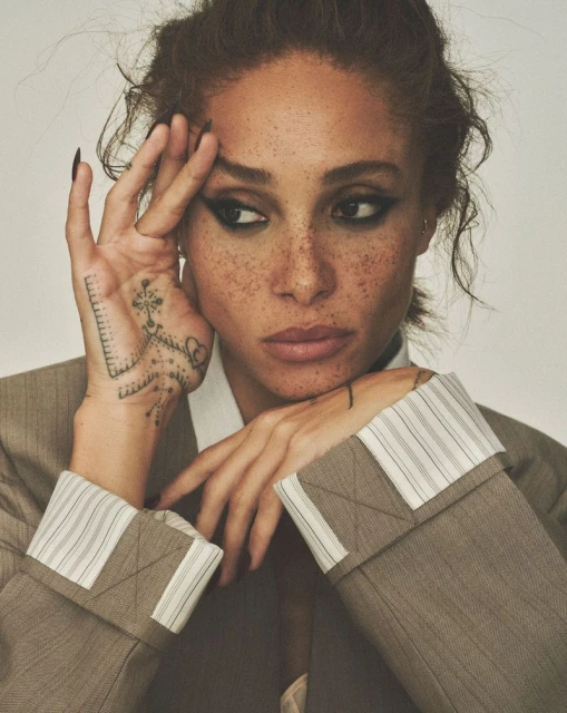 Close-up portrait of an adult woman with freckles and a brown jacket, looking off-camera with hand near her face.