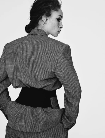 Monochrome studio shot of a stylish adult woman wearing a gray suit with a black waist cincher, looking away from the camera.
