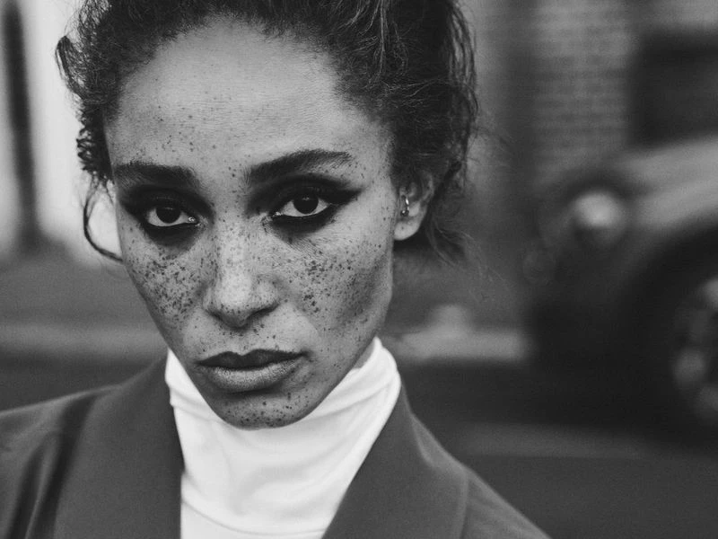 Black and white portrait of a stylish adult woman with freckles wearing a white turtleneck and blazer outdoors in an urban setting.