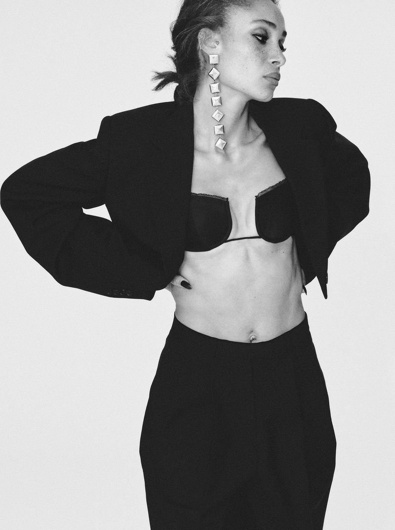 Black and white fashion portrait of an adult female wearing a blazer, bra top, pants, and long earrings against a white backdrop.