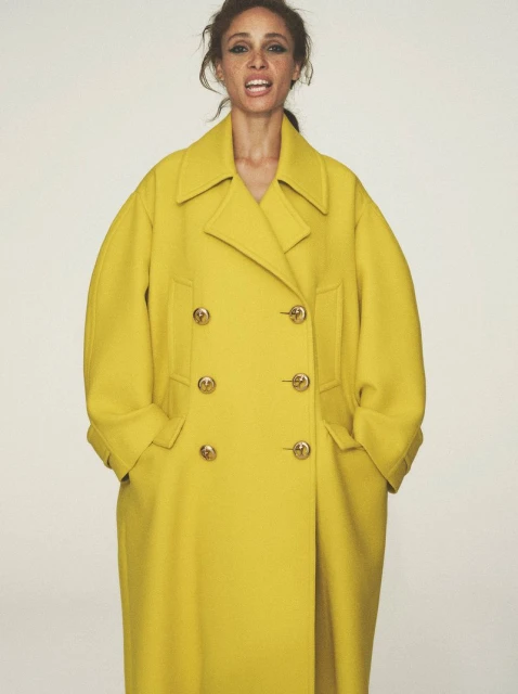 A woman with tan skin wears a bright yellow double-breasted wool coat with gold buttons against a neutral backdrop in a studio shot.