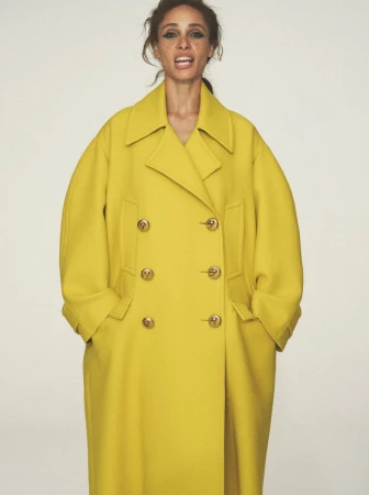 A woman with tan skin wears a bright yellow double-breasted wool coat with gold buttons against a neutral backdrop in a studio shot.