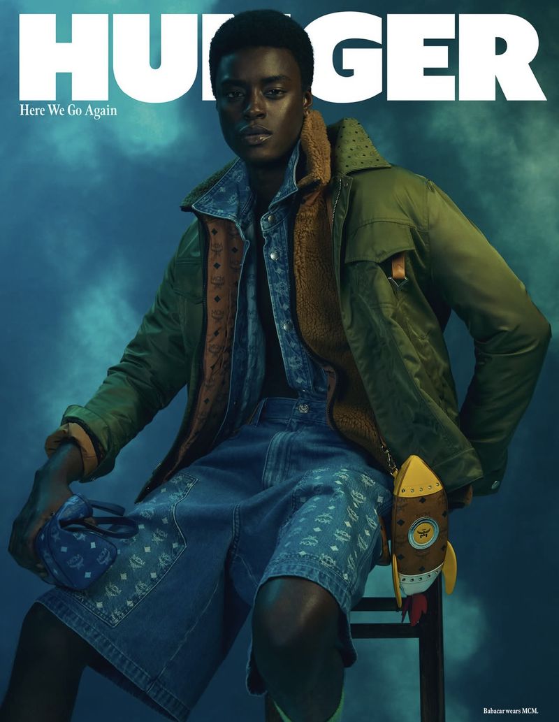 Magazine cover featuring a model in an olive green jacket and denim shorts, holding a blue bag and sitting against a blue background.