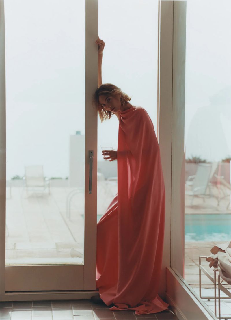 Adult woman in a light pink dress standing by a doorway in an indoor environment with neutral light and tiled floors.