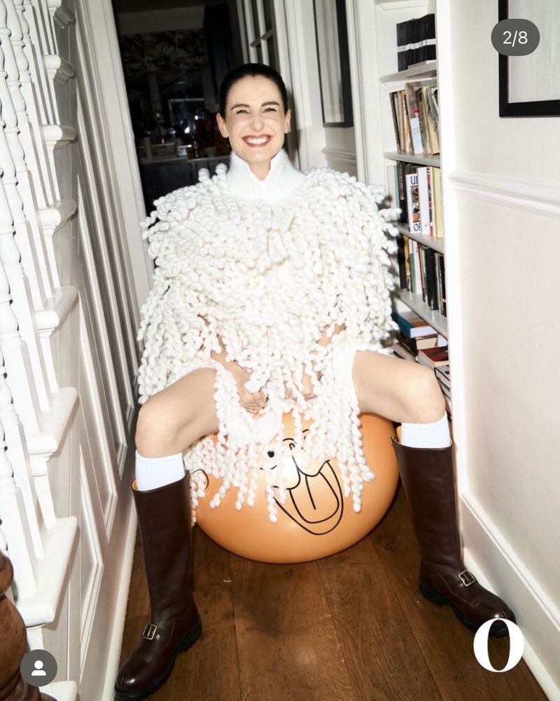 Smiling woman in a white outfit sits on an orange exercise ball indoors next to a staircase and bookshelf.