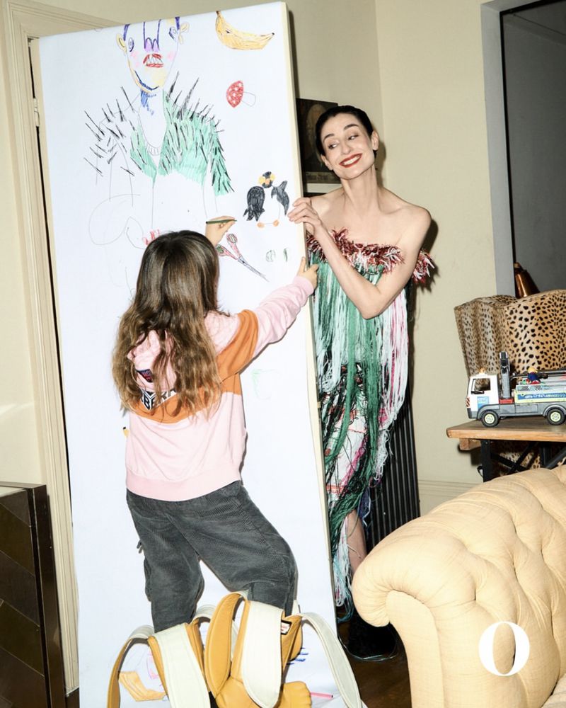 Adult woman and a child stand by a large drawing on canvas in an indoor setting. The woman is wearing a distinctive fringed dress.