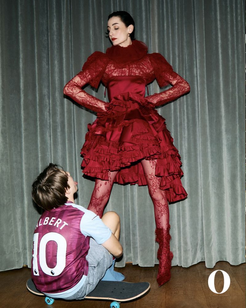 Fashion photo of a woman in an ornate red dress with a boy in a sports jersey sitting on a skateboard in front of her.